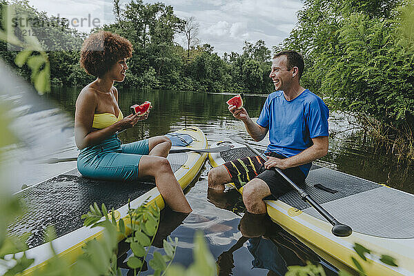 Lächelnde Freunde essen Wassermelone auf dem Paddleboard im See