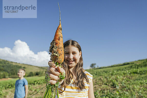 Lächelndes Mädchen zeigt Karotte im Gemüsegarten an einem sonnigen Tag