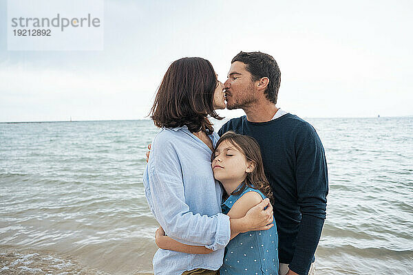 Father and mother kissing each other with daughter in front of sea