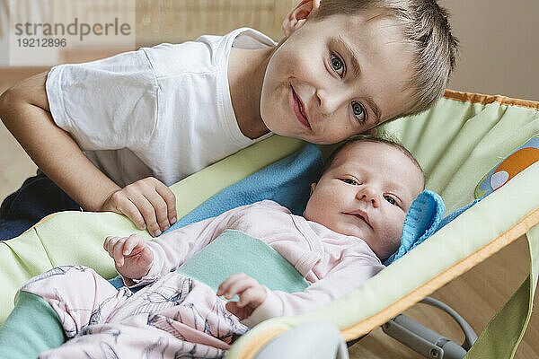 Smiling boy next to newborn sister lying in baby bouncer at home