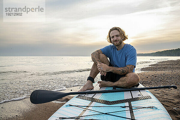 Glücklicher Mann mit Tätowierung sitzt auf einem Paddleboard am Strand