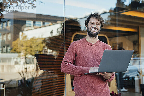 Lächelnder Geschäftsmann mit Laptop vor Glaswand