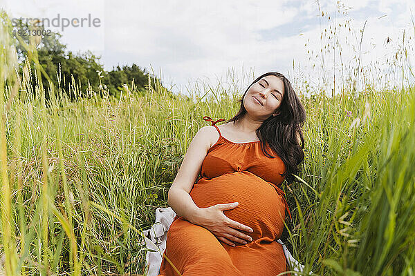 Lächelnde schwangere Frau mit geschlossenen Augen sitzt inmitten von Gras
