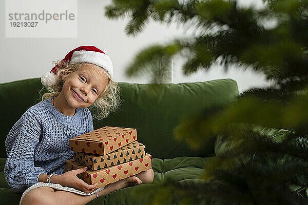 Lächelndes Mädchen mit Weihnachtsgeschenken sitzt zu Hause auf dem Sofa