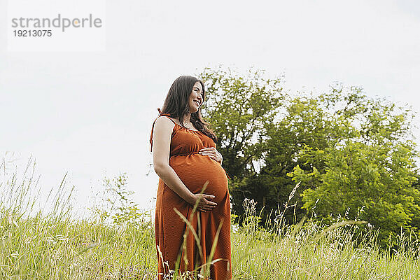 Smiling pregnant woman with hands on stomach standing amidst grass