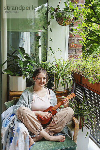 Lächelndes Teenager-Mädchen mit Ukulele sitzt auf einem Stuhl auf dem Balkon