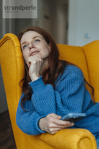 Nachdenkliche Frau sitzt mit Smartphone auf gelbem Sessel zu Hause