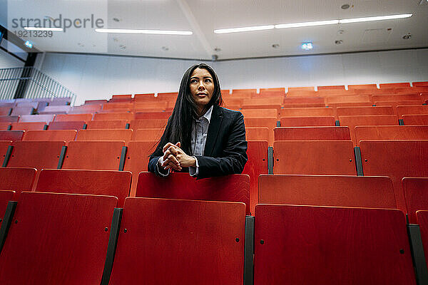 Confident businesswoman with hands clasped leaning on red seats in auditorium