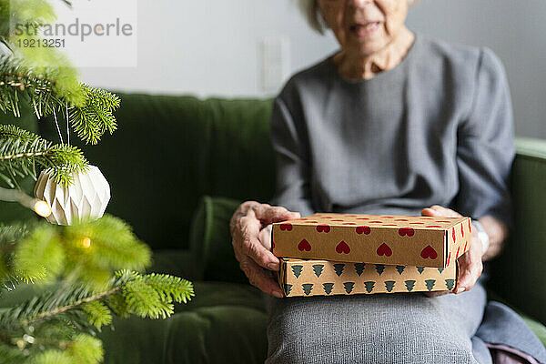 Hände einer älteren Frau  die Geschenke in der Hand hält und zu Hause neben dem Weihnachtsbaum auf dem Sofa sitzt