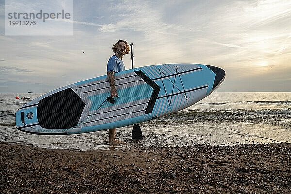 Junger Mann trägt Paddleboard am Strand