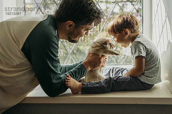 Father giving cute mixed breed to son sitting in window at home