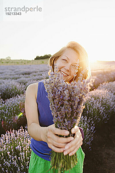 Smiling woman holding bunch of lavender flowers in field on sunny day