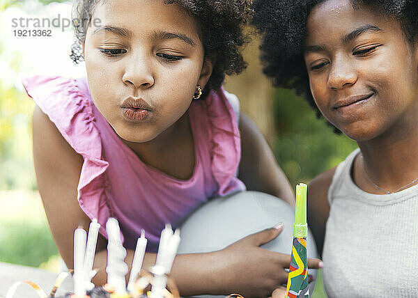 Lächelndes Mädchen mit Schwester bläst Geburtstagskerzen im Park