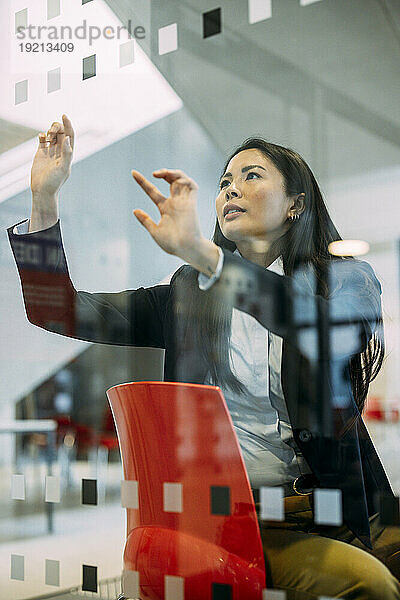 Selbstbewusste Geschäftsfrau sitzt auf einem Stuhl und berührt die Glaswand am Arbeitsplatz