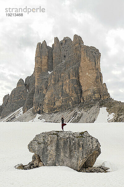 Man practicing yoga on rock surrounded by snow in front of mountain