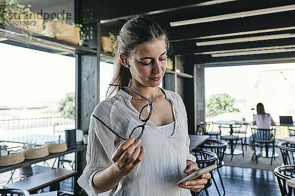 Portrait of pretty blonde standing in cafe with eyeglasses and smart phone in hands