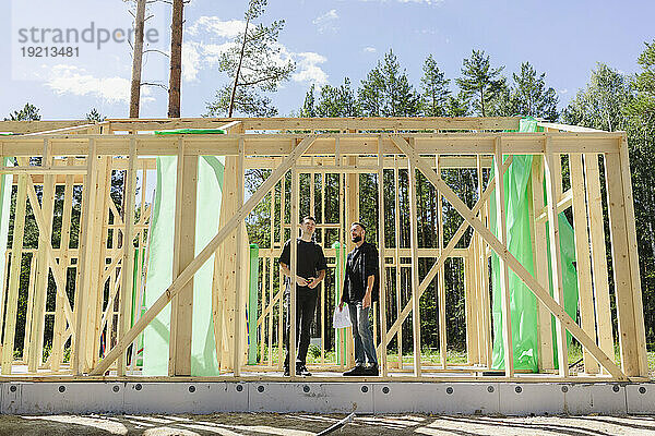 Ingenieure stehen an einem sonnigen Tag mit Bauplänen in der Nähe eines Holzrahmens