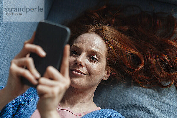 Lächelnde rothaarige Frau benutzt Smartphone auf dem Sofa