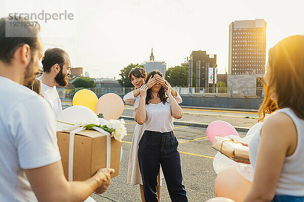 Freunde überraschen Frau mit Luftballons und Geschenkbox bei Sonnenuntergang
