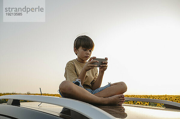 Junge sitzt auf Autodach und benutzt Smartphone