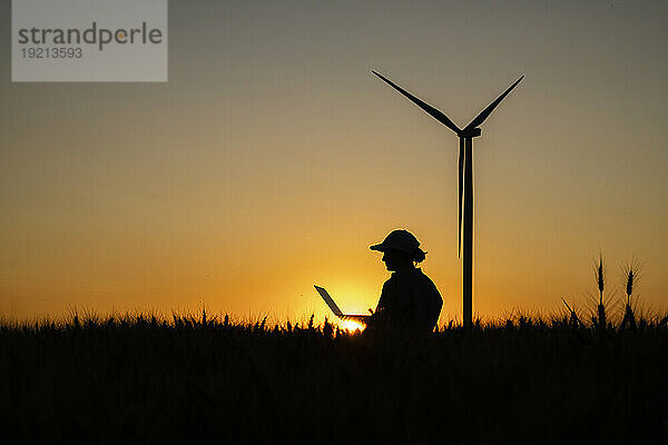 Silhouette eines Bauern  der in der Abenddämmerung einen Laptop auf einem Feld mit einer Windkraftanlage benutzt