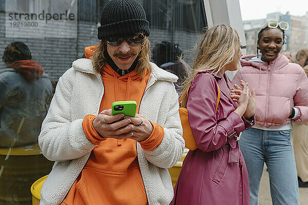 Lächelnder Mann benutzt Smartphone mit Freunden im Hintergrund in der Nähe einer Glaswand