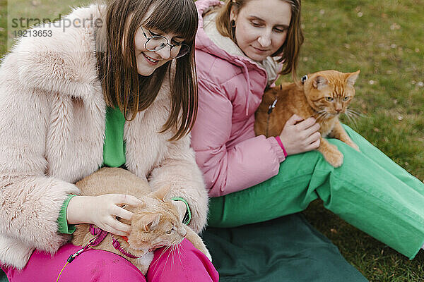 Zwei junge Frauen halten Katzen und sitzen auf Gras im Park