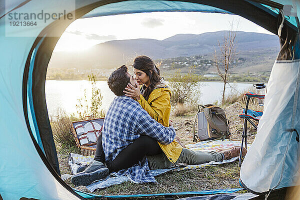 Affectionate couple sitting on picnic blanket near tent