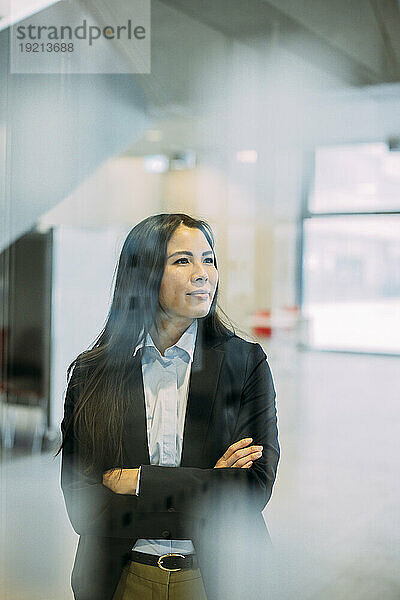 Businesswoman standing with arms crossed seen through glass at workplace