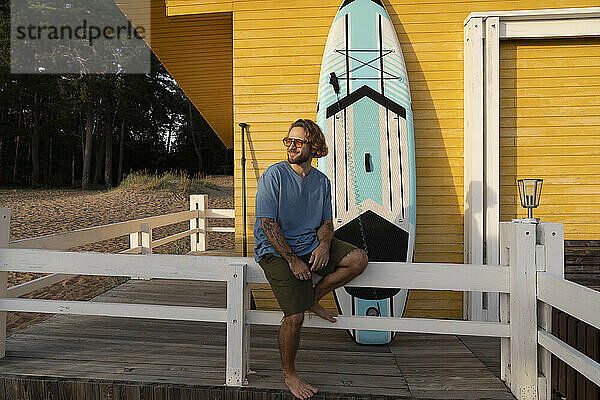 Smiling man sitting near paddleboard in front of beach hut