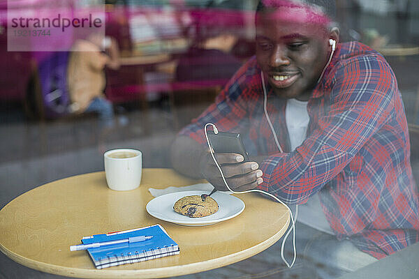 Lächelnder junger Mann  der im Café ein Smartphone benutzt und Musik hört