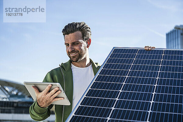 Lächelnder Ingenieur hält an einem sonnigen Tag ein digitales Tablet und ein Solarpanel in der Hand