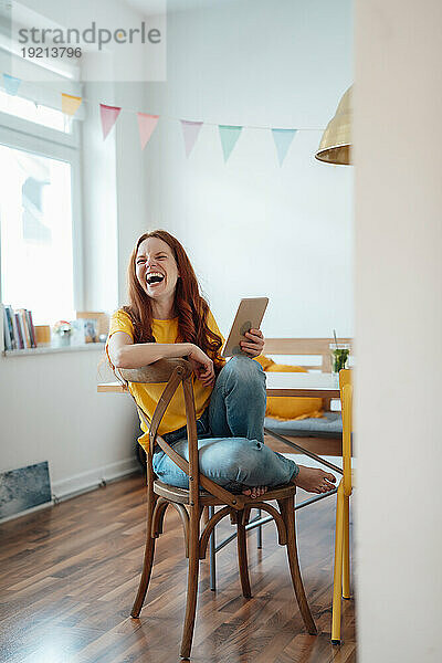 Fröhliche Frau lacht und sitzt zu Hause mit Tablet-PC auf einem Stuhl