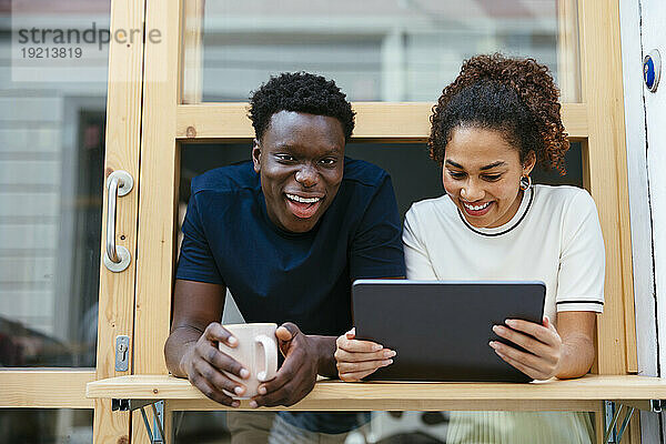 Lächelnder junger Student mit Freund  der Tablet-PC benutzt und sich aus dem Fenster lehnt