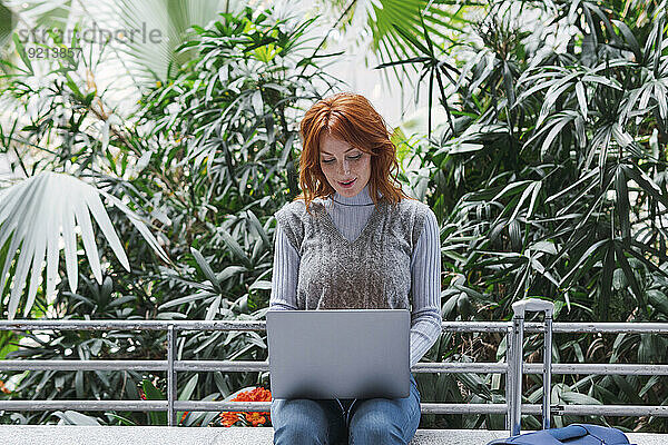 Lächelnde rothaarige Geschäftsfrau mit Laptop vor Pflanzen