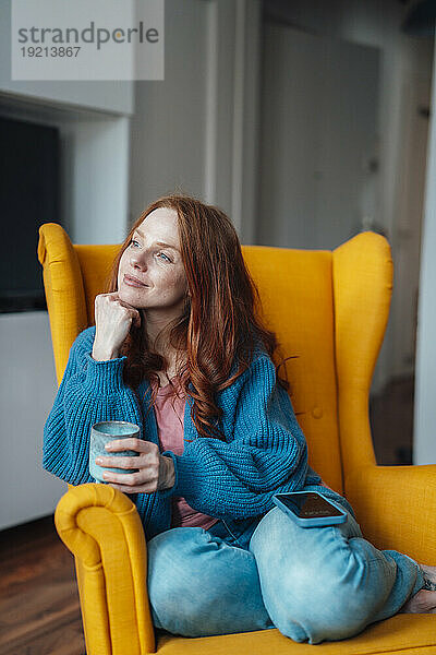 Nachdenkliche Frau sitzt im Sessel mit Kaffeetasse und Smartphone
