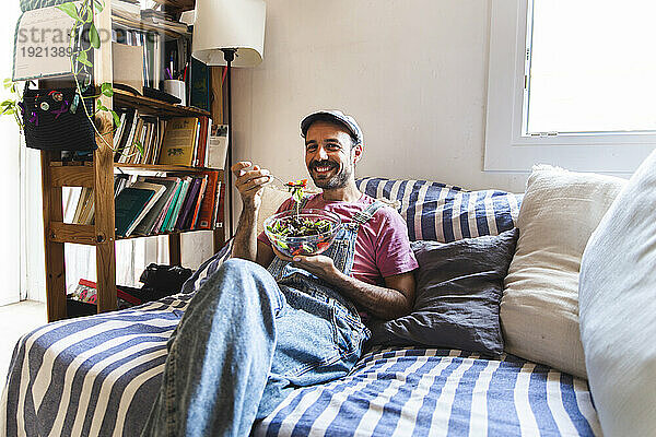 Lächelnder Mann entspannt sich und isst Salat auf dem Sofa zu Hause