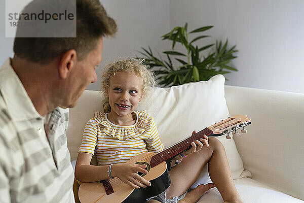 Lächelndes Mädchen  das Gitarre spielt  sitzt neben seinem Vater im Wohnzimmer zu Hause