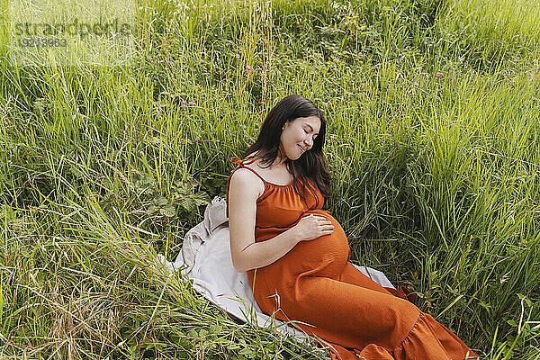 Pregnant woman sitting on blanket near grass in meadow