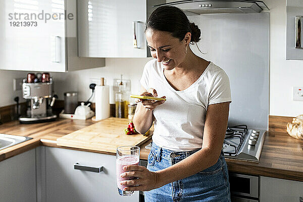 Glückliche Frau fotografiert Milchshake per Smartphone in der heimischen Küche