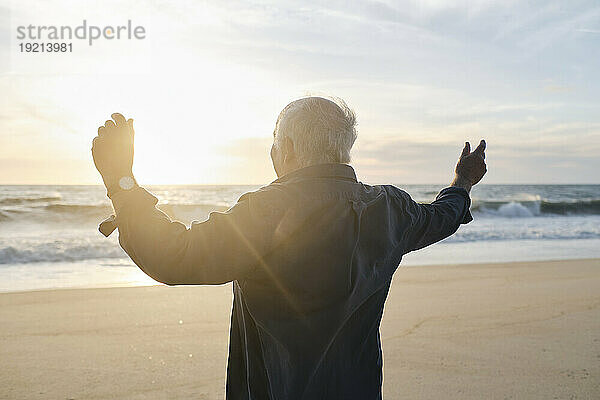 Älterer Mann mit erhobenen Armen steht vor dem Meer am Strand