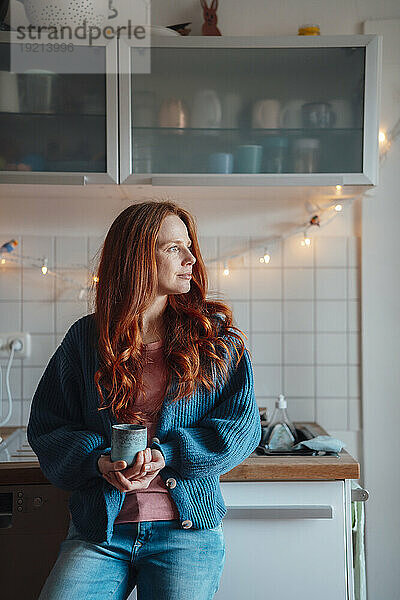 Nachdenkliche Frau steht mit Kaffeetasse in der heimischen Küche