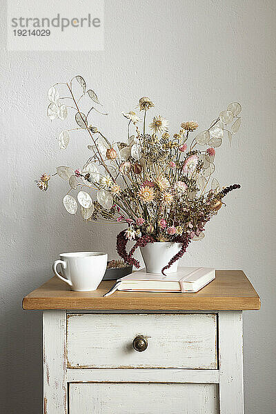 Note pad  mug and arrangement of dried flowers on small cabinet