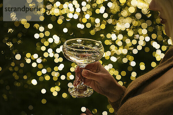 Frau hält Champagnerglas neben beleuchteten Weihnachtslichtern