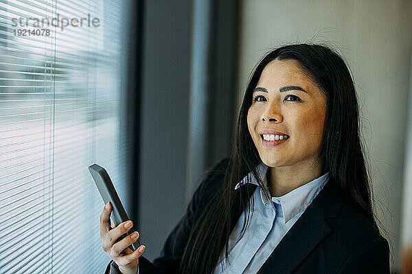 Lächelnde Geschäftsfrau benutzt Smartphone am Fenster im Büro