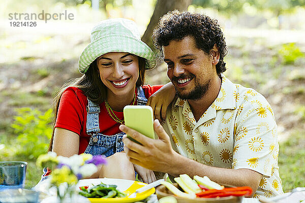 Lächelnder Mann zeigt einem Freund am Picknicktisch sein Smartphone