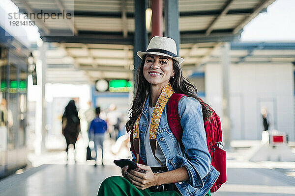 Glückliche Frau mit Rucksack und Smartphone sitzt an der Straßenbahnhaltestelle