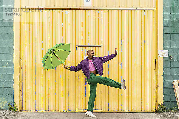 Nicht-binäre Person tanzt mit grünem Regenschirm vor gelber Fensterladentür