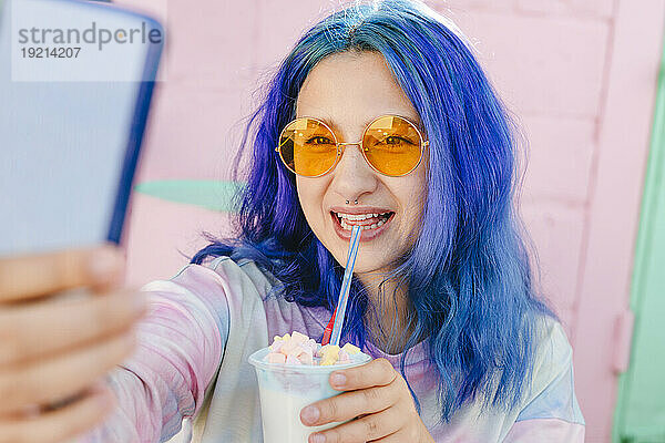 Glückliche Frau mit gefärbten blauen Haaren hält ein Getränk in der Hand und macht ein Selfie im Café