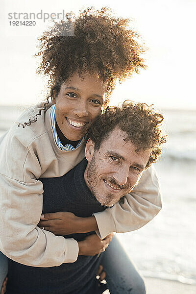 Lächelnder Mann huckepack mit seiner Freundin am Strand